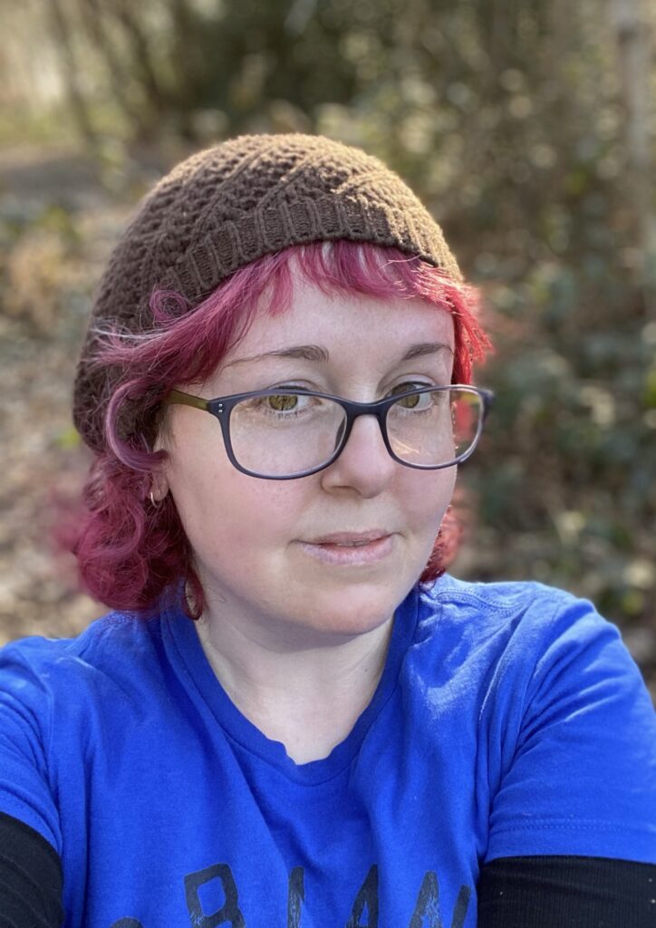 Ro Stark, a white person, with dyed red hair and glasses, smiling at the camera. 